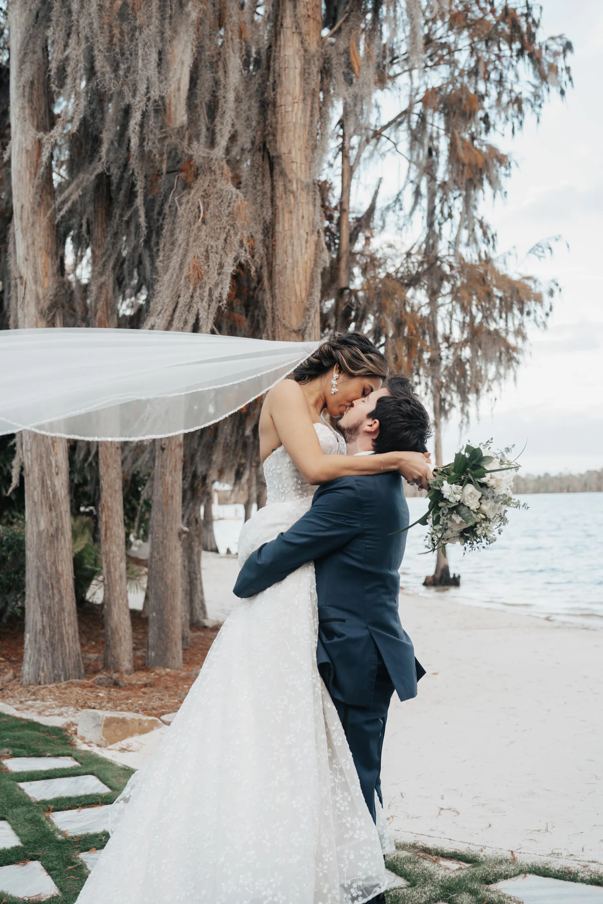 A groom lifts his bride for a kiss at the water’s edge at Paradise Cove on Lake Bryan, her veil streaming in the wind under moss-draped cypress trees
