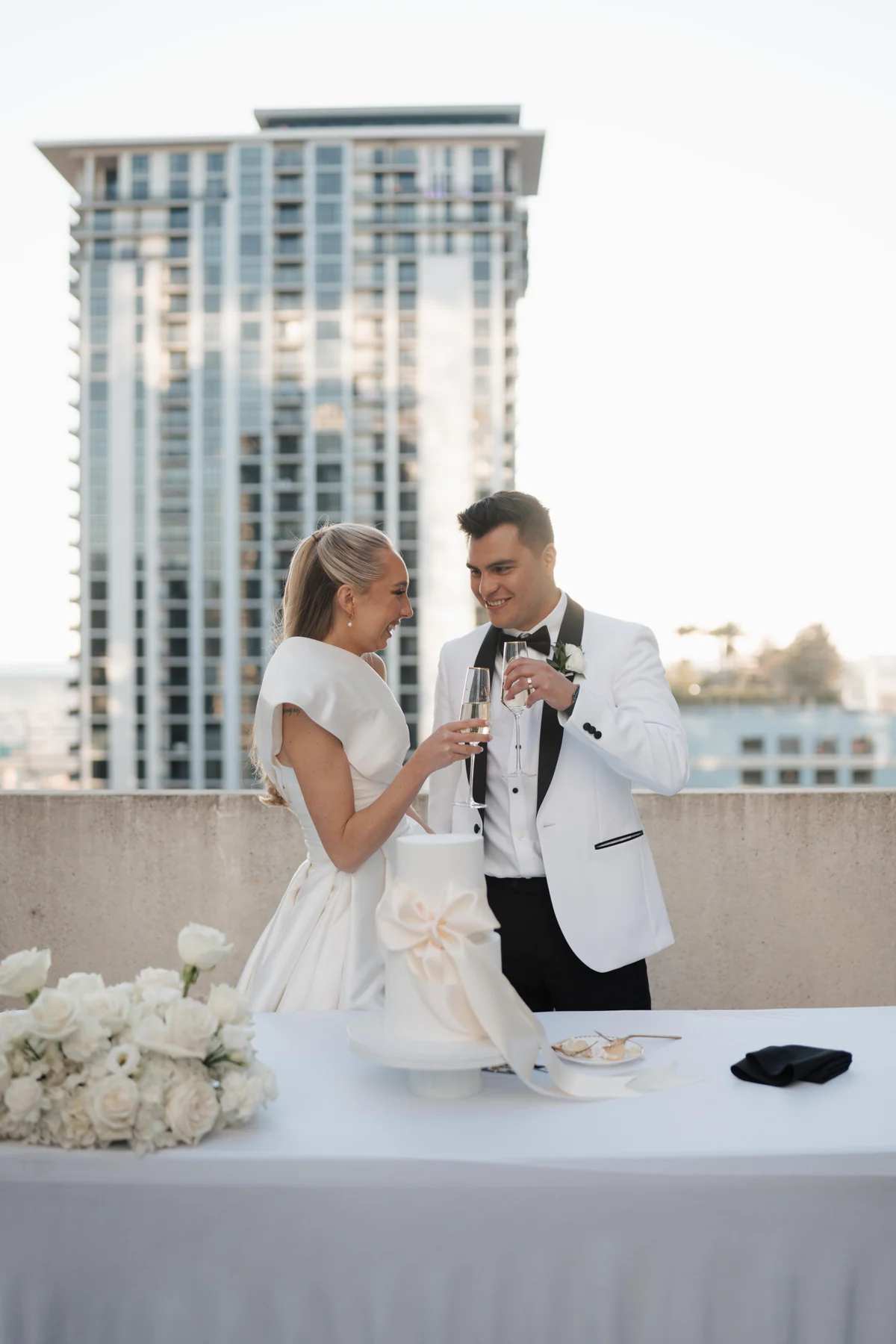 A newly married couple toasting with champagne beside their wedding cake on a rooftop