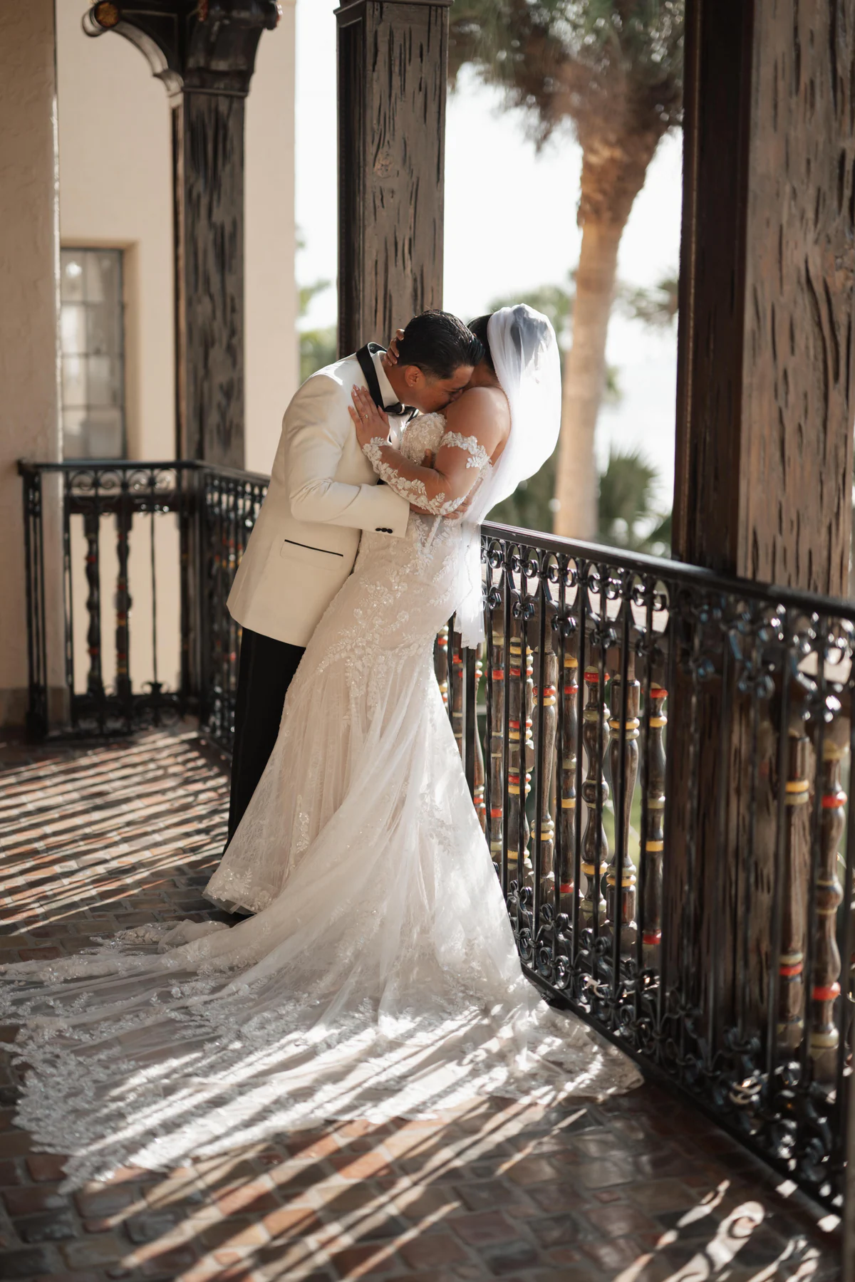 Amanda and Sebastian embracing on the balcony at Powel Crosley Estate in Sarasota, with the iron railing, palm trees, and afternoon sun behind them