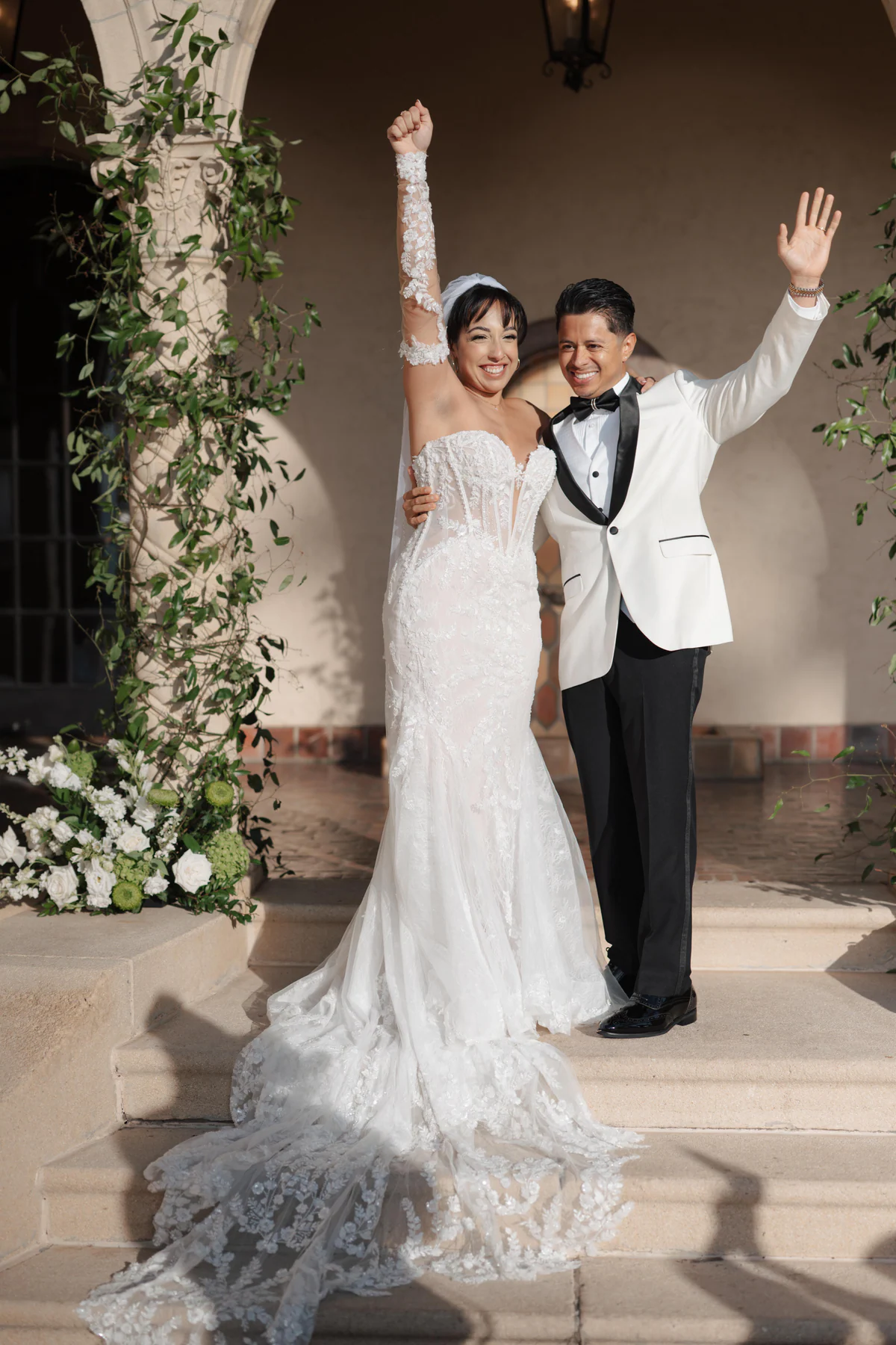 Amanda and Sebastian celebrating with raised arms on the steps after their ceremony at Powel Crosley Estate in Sarasota, with white flowers and greenery beside them