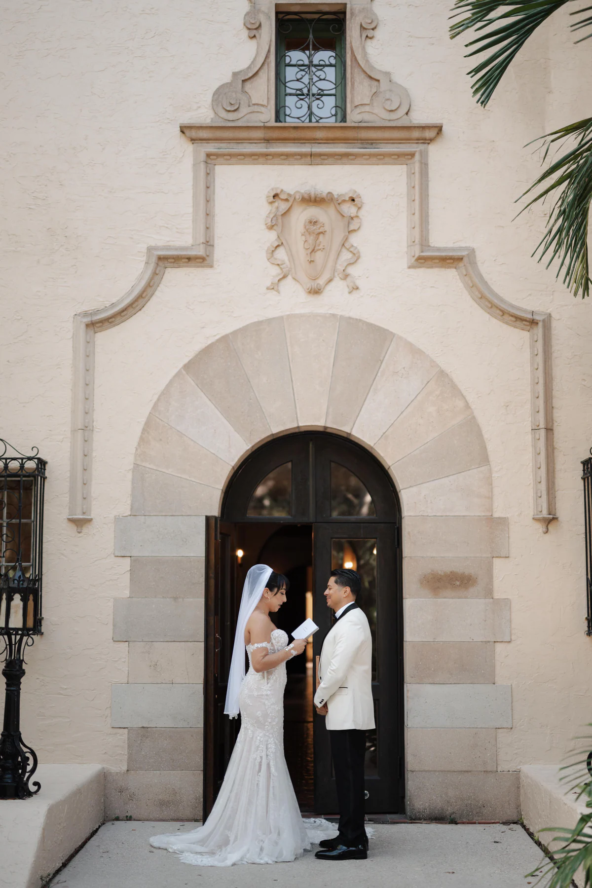 Amanda and Sebastian reading their vows together at the arched entrance of Powel Crosley Estate in Sarasota, with the mansion facade and carved crest behind them