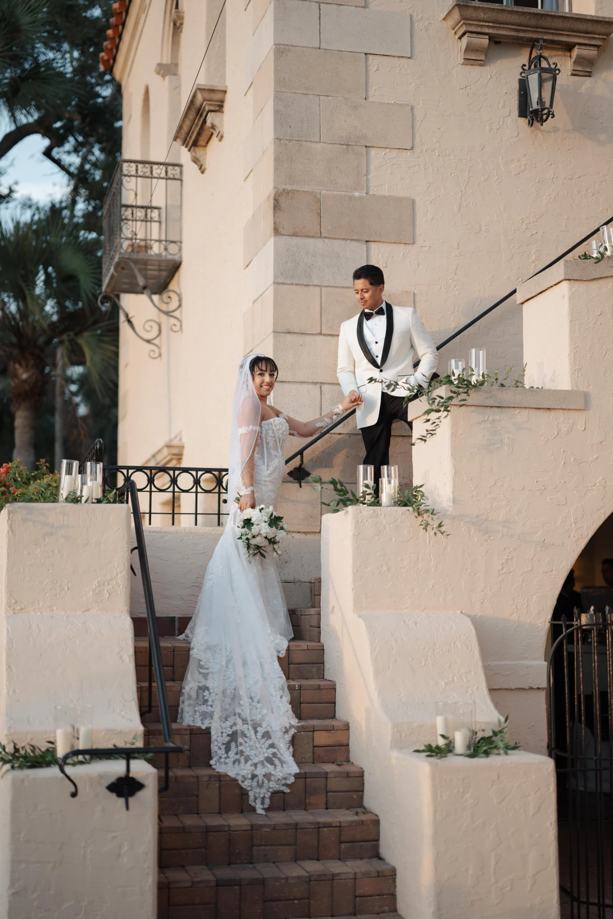 Amanda and Sebastian holding hands on the exterior stairs of Powel Crosley Estate in Sarasota, with candles, greenery, and the mansion wall behind them