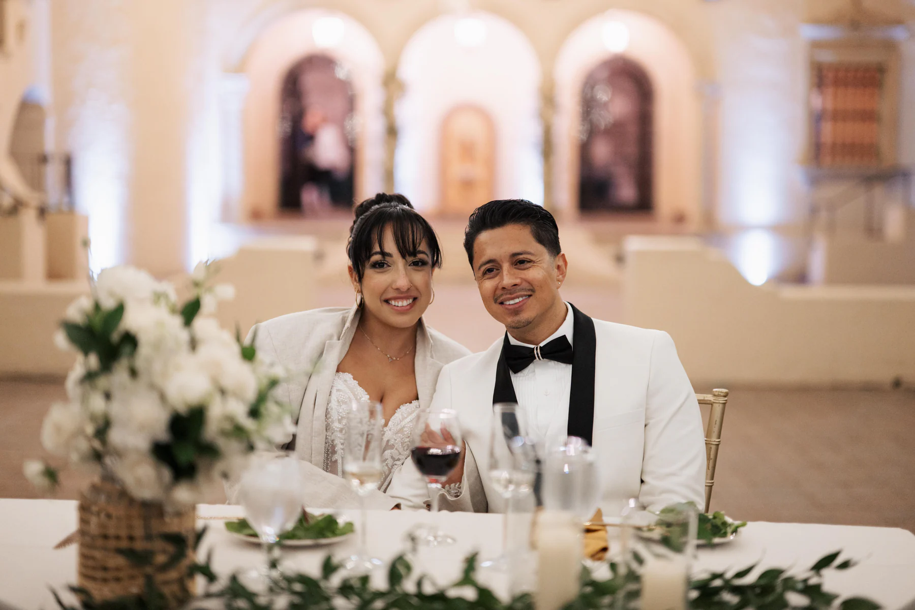 Amanda and Sebastian smiling together at their reception table at Powel Crosley Estate in Sarasota, with white flowers, candles, and the arched reception room behind them